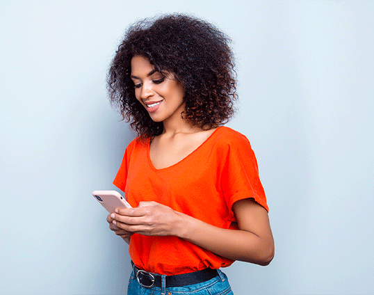 Mujer de cabello riso y camisa anaranjada sonriendo mientras observa su dispositivo móvil 